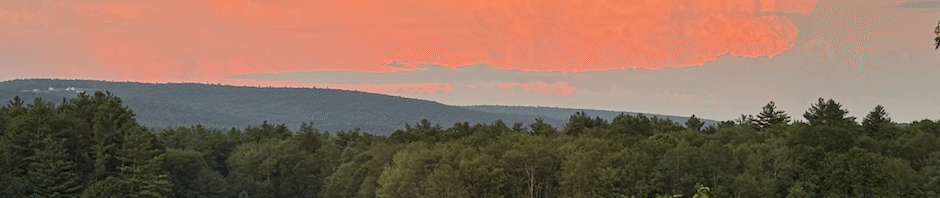 Sunrise over woods, with sky showing mostly a bright tangerine cloud against a small amount of gray sky in top half of photo. Bottom half shows very dark wooded hills, with tops of trees, dark green, in foreground.